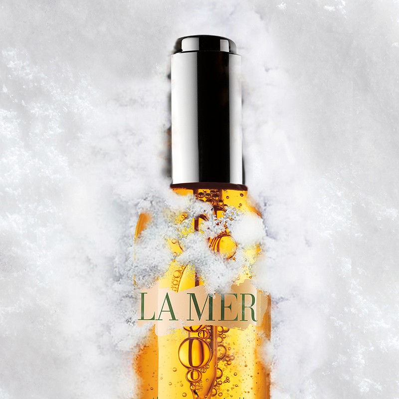 La Mer face oil in clear bottle with silver cap surrounded by snow. Studio shot with white background. Beauty product.