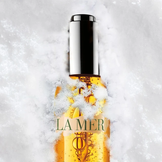 La Mer face oil in clear bottle with silver cap surrounded by snow. Studio shot with white background. Beauty product.