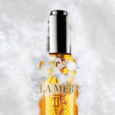 La Mer face oil in clear bottle with silver cap surrounded by snow. Studio shot with white background. Beauty product.