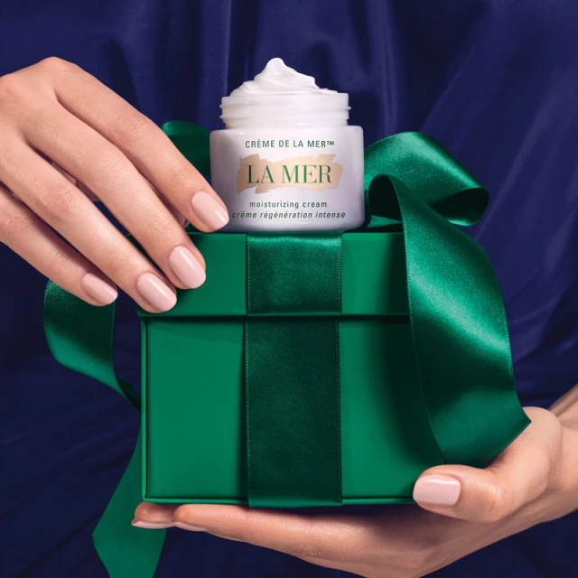 A person holds a green gift box with a La Mer moisturizing cream product placed on top, set against a navy blue fabric backdrop.