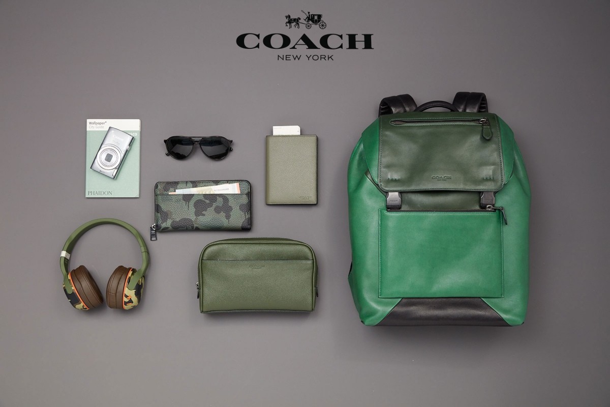 Flat lay studio shot of a Coach green and black leather backpack, wallet, pouch, sunglasses, and headphones on a grey background.