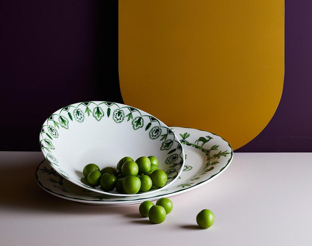 Still life of two stacked white plates with green floral and deer designs filled with green plums, against a purple and yellow backdrop.