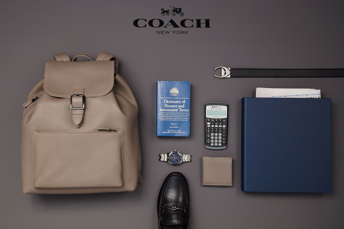 Beige leather Coach backpack surrounded by a finance book, calculator, wallet, watch, shoe, belt, and folder on a grey background.