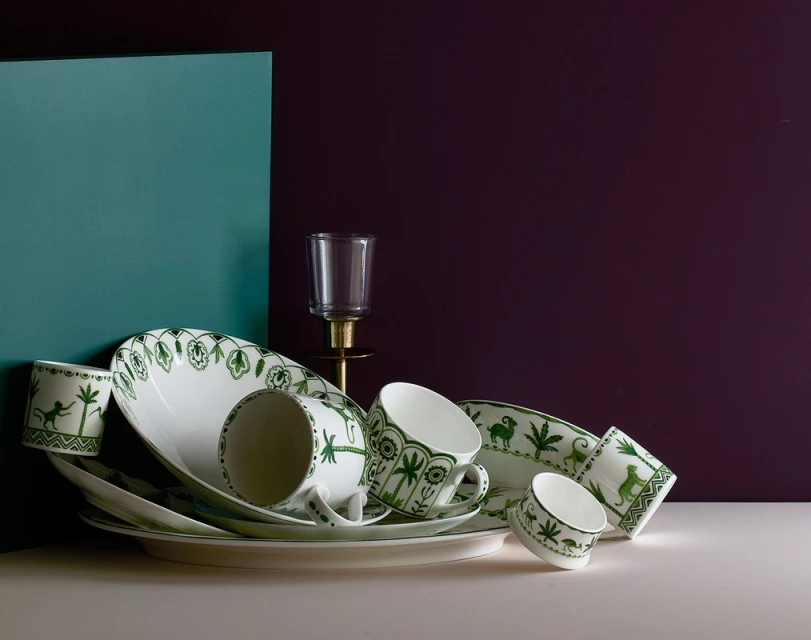 Arrangement of white ceramic tableware with green foliage and animal pattern designs, set against teal and dark purple backdrop.