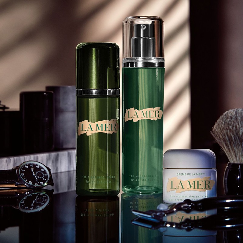 La Mer skincare product bottles and jar on a reflective surface, showcasing the brand's collection with shaving brush and razor.