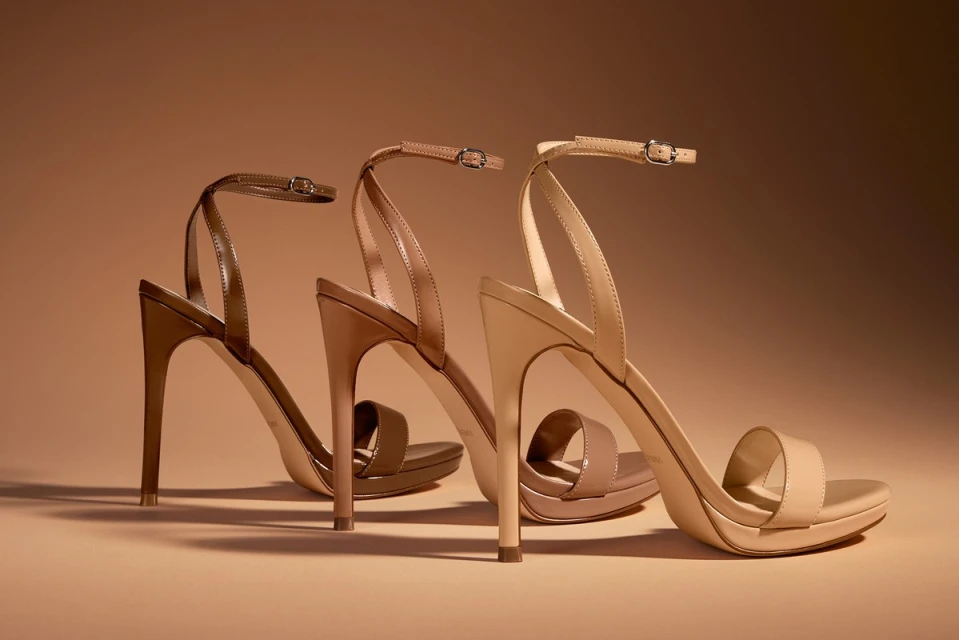 Studio shot of three high-heel shoes, each a different shade of tan and brown, arranged against a light brown backdrop.