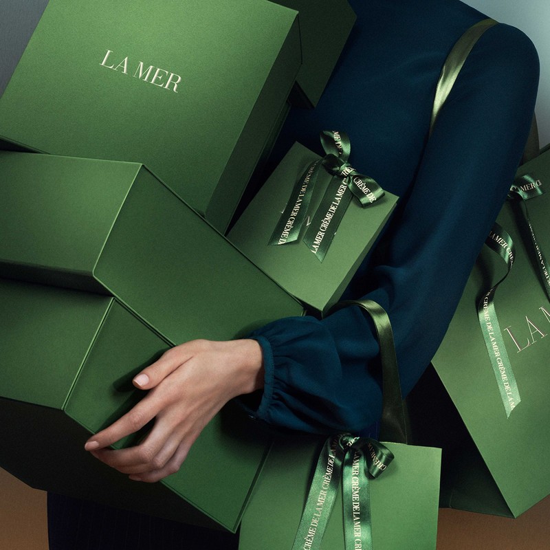 Woman holding stacked green La Mer skincare gift boxes with ribbons in studio setting promoting beauty and skincare products.
