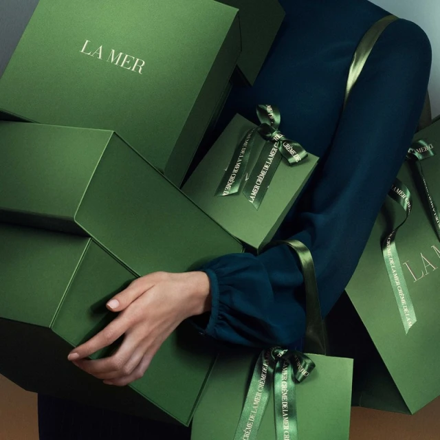 Woman holding stacked green La Mer skincare gift boxes with ribbons in studio setting promoting beauty and skincare products.