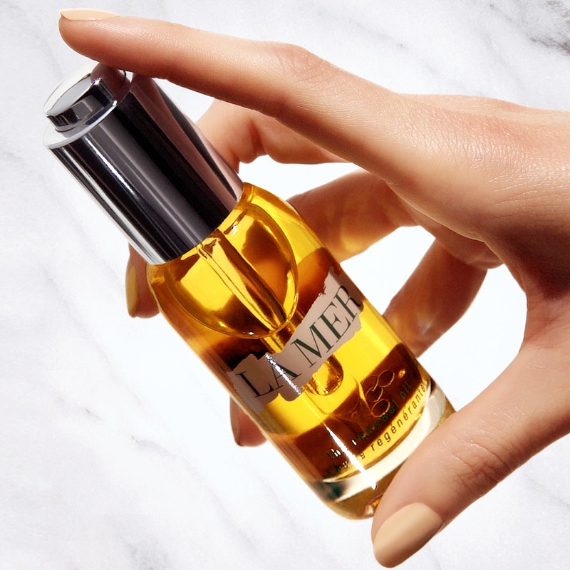 A hand holds a La Mer facial oil bottle against a marble background, showcasing the product's luxurious packaging and golden oil.