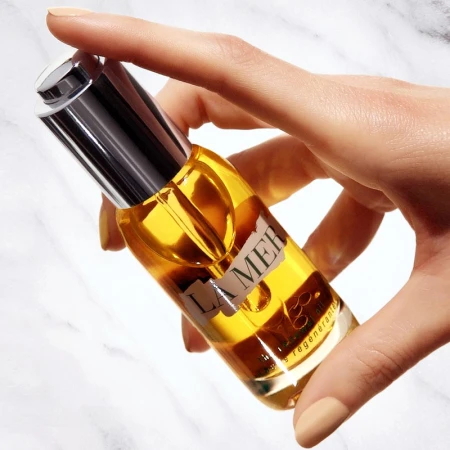 A hand holds a La Mer facial oil bottle against a marble background, showcasing the product's luxurious packaging and golden oil.