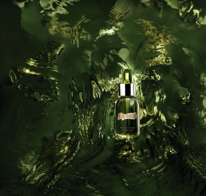 La Mer renewal oil sits in the center of a green reflective pool, reflecting light. The bottle has a green dropper top.