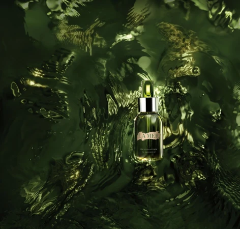 La Mer skincare bottle resting vertically on shimmering green rippling liquid surface with metallic highlights