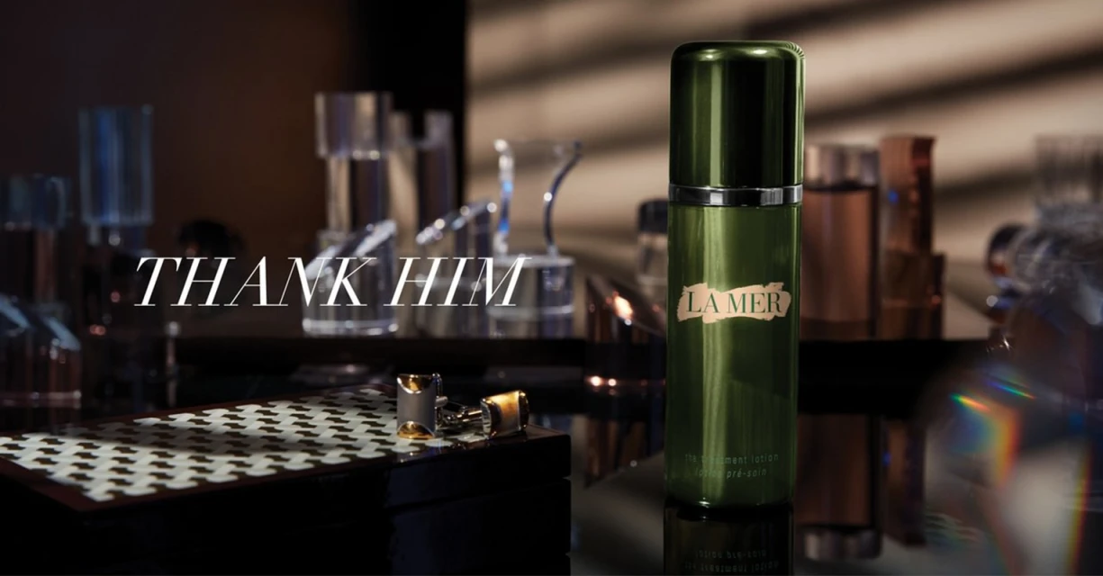La Mer treatment lotion and cufflinks on a reflective dark surface, clear vases, and text 'Thank Him' in a beauty advertisement.