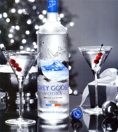 Grey Goose vodka bottle displayed with martini glasses garnished with cherries, set against a blurred, festive background.