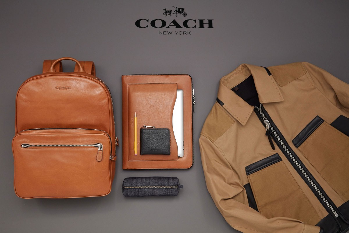 Flat lay studio shot of a leather backpack, jacket, notebook, pencil, wallet, and pencil case against a neutral gray backdrop.