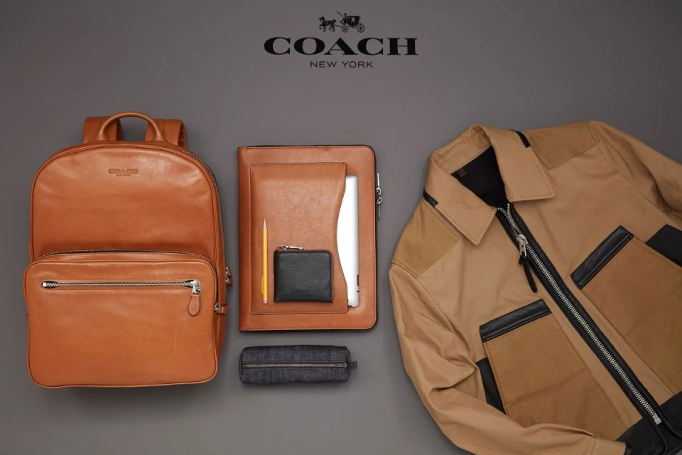 Flat lay studio shot of a leather backpack, jacket, notebook, pencil, wallet, and pencil case against a neutral gray backdrop.