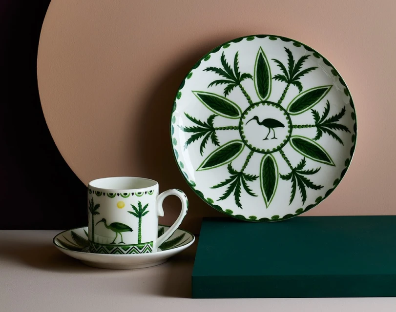 Stylized product shot: white ceramic plate and cup with green tropical foliage and heron design against a neutral background.