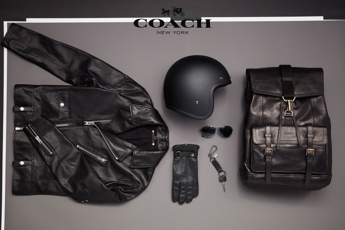 Flat lay studio shot of black leather biker jacket, backpack, helmet, gloves, sunglasses and keychain on grey background.