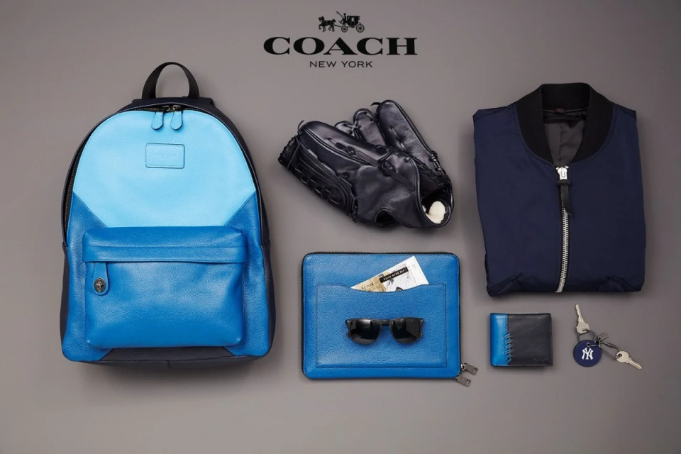 Coach brand product shot: blue leather backpack, baseball glove, jacket, clutch, sunglasses, wallet, and New York Yankees keychain.