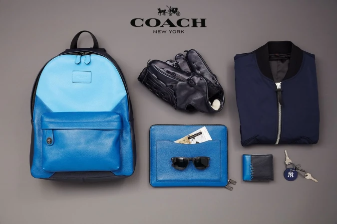 Coach brand product shot: blue leather backpack, baseball glove, jacket, clutch, sunglasses, wallet, and New York Yankees keychain.