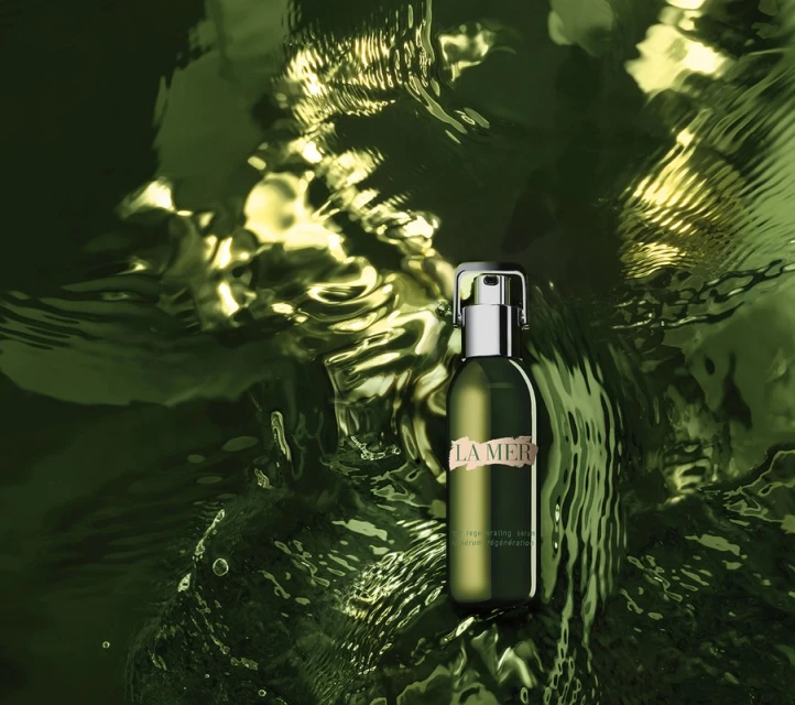 La Mer regenerating serum in a green bottle with a silver pump top is surrounded by water, creating a luxurious beauty product shot.