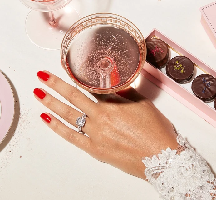 Close-up shot of a woman's hand adorned with a diamond ring, holding a champagne glass, with chocolates in the background.