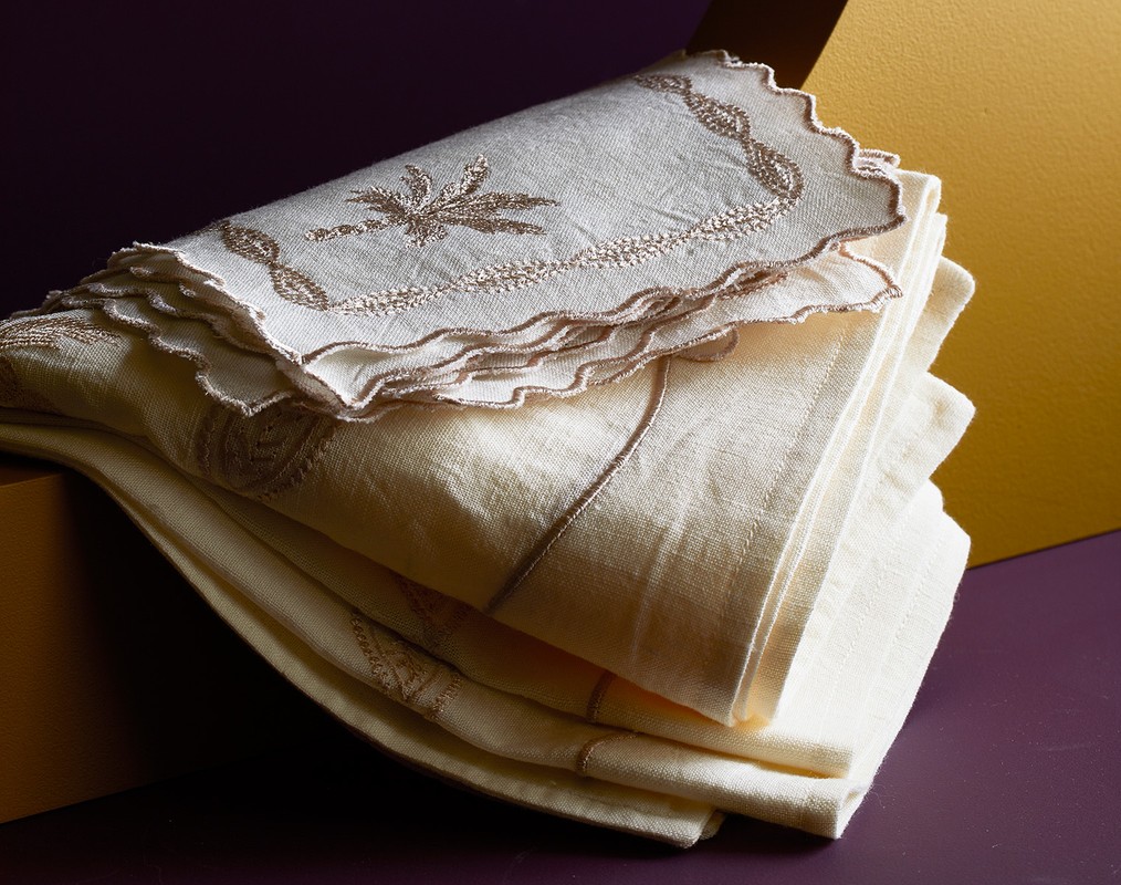Close-up shot of a neatly stacked pile of embroidered, off-white linen napkins with decorative edges, set against a purple and gold background.