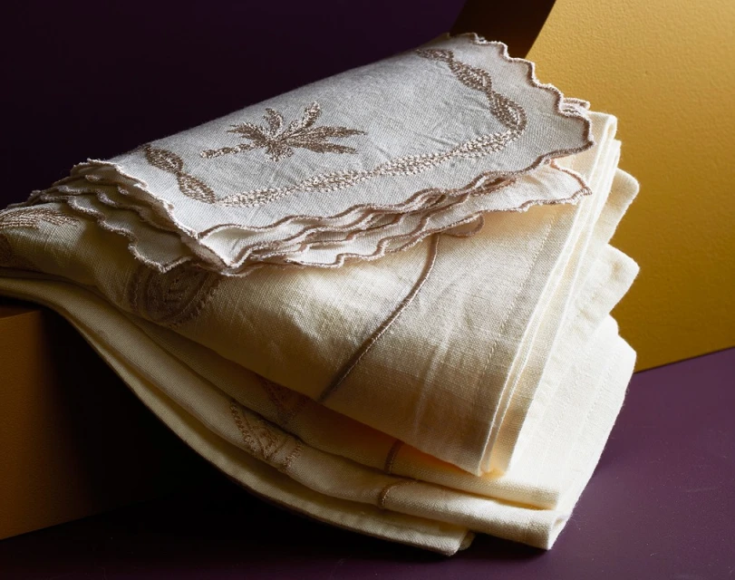 Close-up shot of a neatly stacked pile of embroidered, off-white linen napkins with decorative edges, set against a purple and gold background.