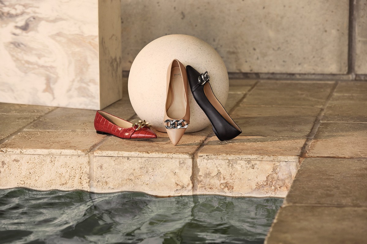 Elegant footwear presentation: Three women's ballet flats in red, beige, and black with chain accents, arranged on a tile surface near water.