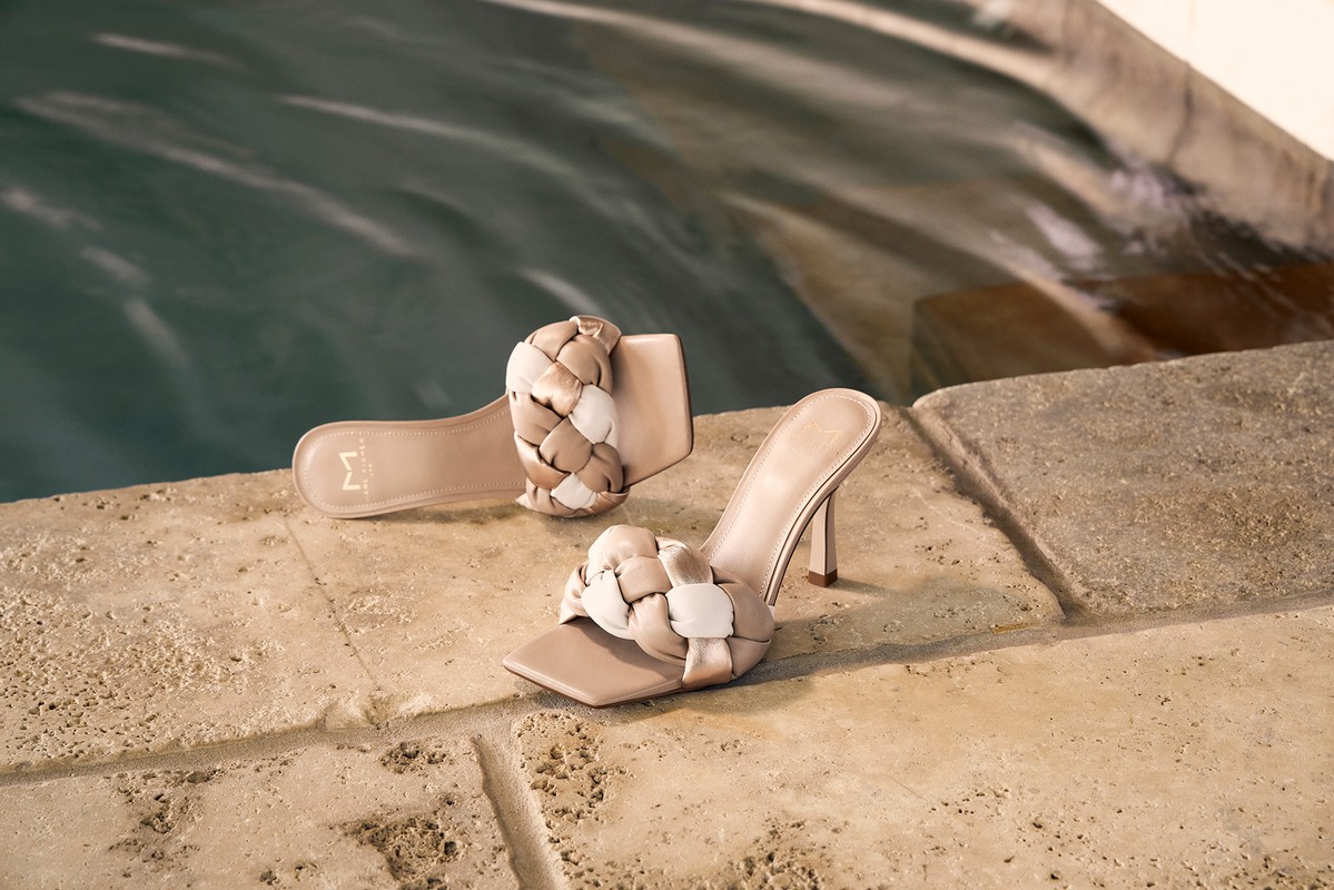 Elegant beige high-heel shoes with braided straps are arranged near a water feature on a stone surface, illuminated by soft, natural light.