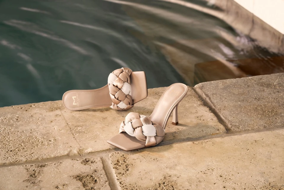 Elegant beige high-heel shoes with braided straps are arranged near a water feature on a stone surface, illuminated by soft, natural light.