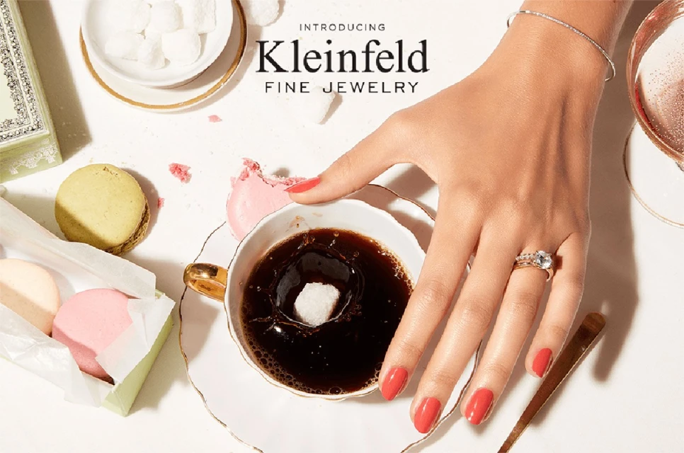 Close-up of woman's hand wearing jewelry, dropping sugar cube into coffee cup, macarons, and luxury branding; overhead shot.