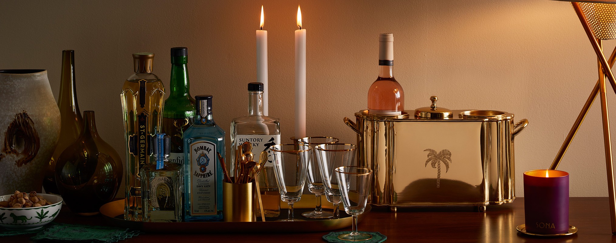 Still life of assorted liquors, glasses, ice bucket, candles, and table lamp on a wooden bar top with a beige wall backdrop.