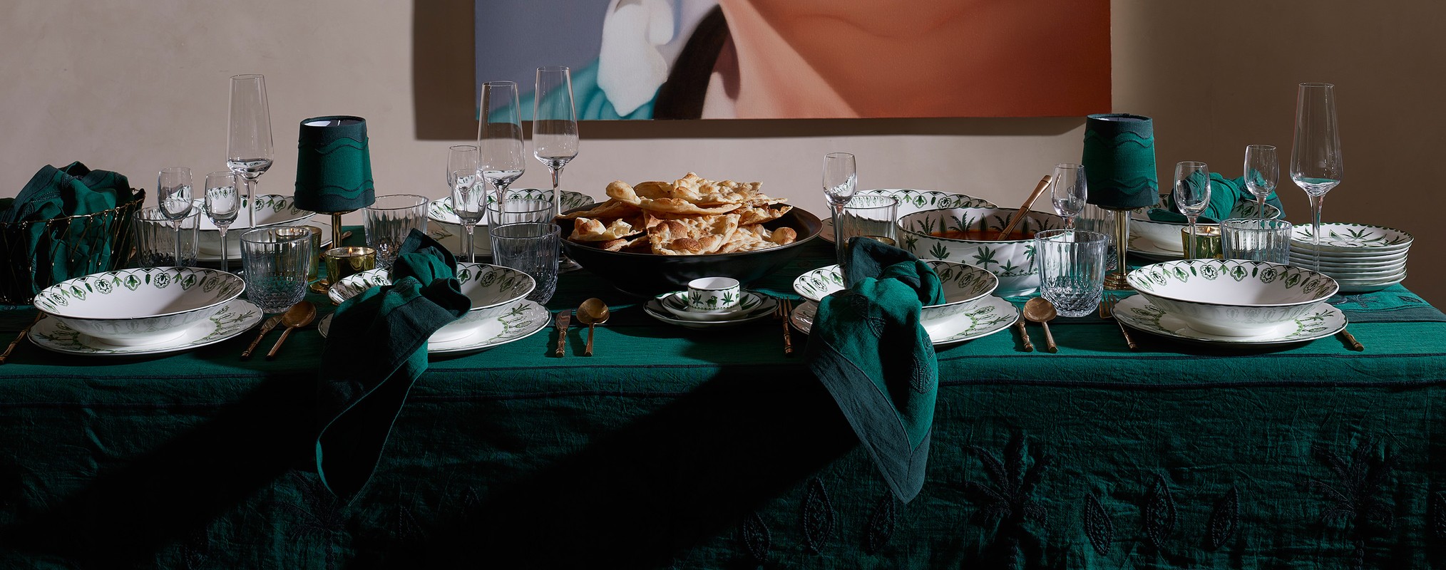 Opulent tablescape featuring ornate white and green patterned dinnerware, crystal glassware, gold cutlery, and dark green linen on a green tablecloth.