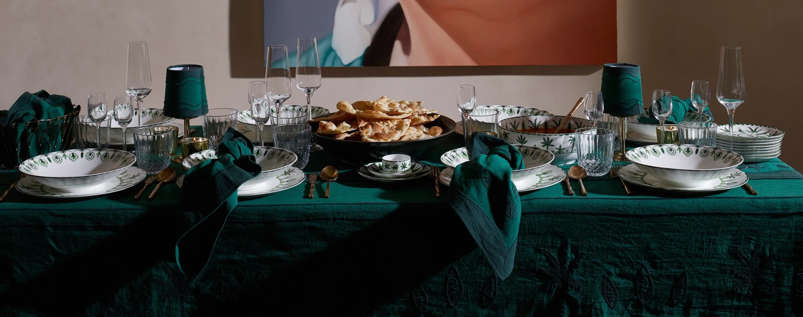 Opulent tablescape featuring ornate white and green patterned dinnerware, crystal glassware, gold cutlery, and dark green linen on a green tablecloth.