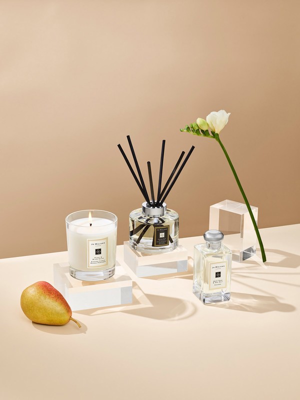 Still life of a candle, fragrance diffuser, perfume bottle, pear, and white flower, presented on clear blocks with a beige backdrop.