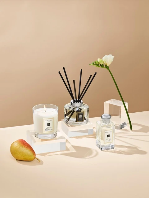 Still life of a candle, fragrance diffuser, perfume bottle, pear, and white flower, presented on clear blocks with a beige backdrop.
