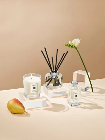 Still life of a candle, fragrance diffuser, perfume bottle, pear, and white flower, presented on clear blocks with a beige backdrop.