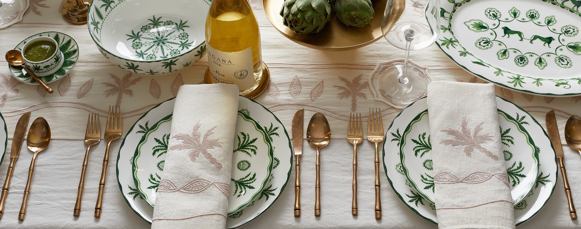 Elegant table setting with white and green patterned plates, linen napkins, gold cutlery, wine bottle, artichokes, and palm tree accents.