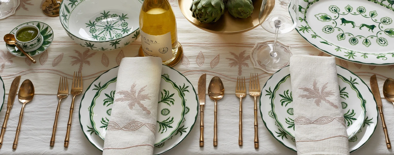 Elegant table setting with white and green patterned plates, linen napkins, gold cutlery, wine bottle, artichokes, and palm tree accents.