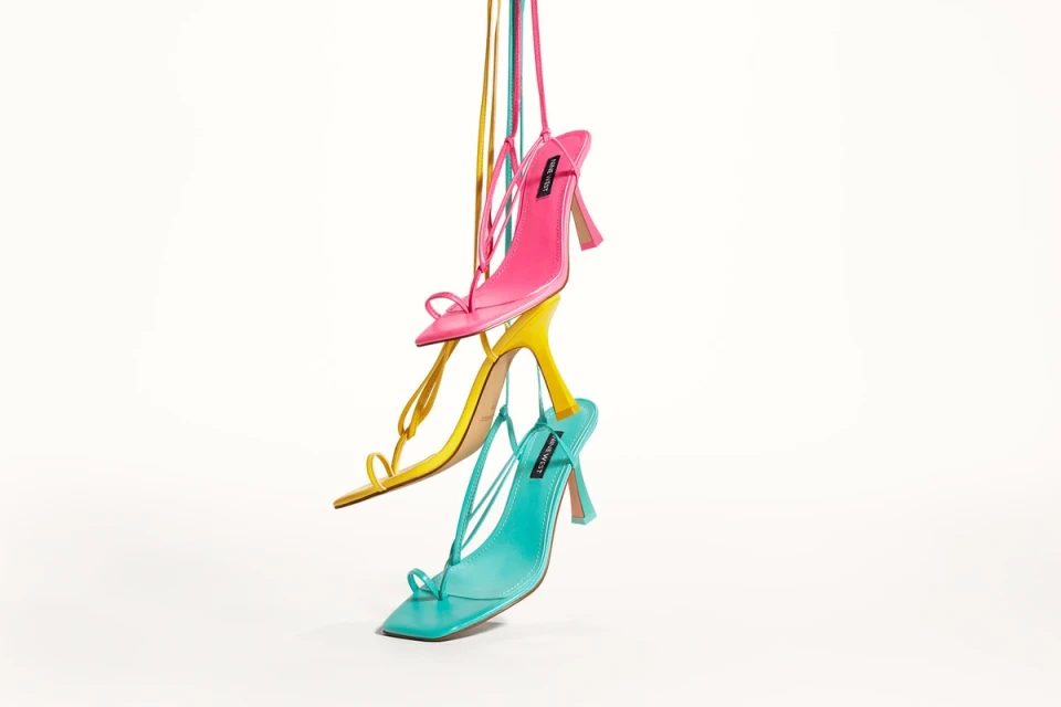 Three brightly colored heeled sandals, pink, yellow, and turquoise, stacked and suspended against a stark white backdrop.