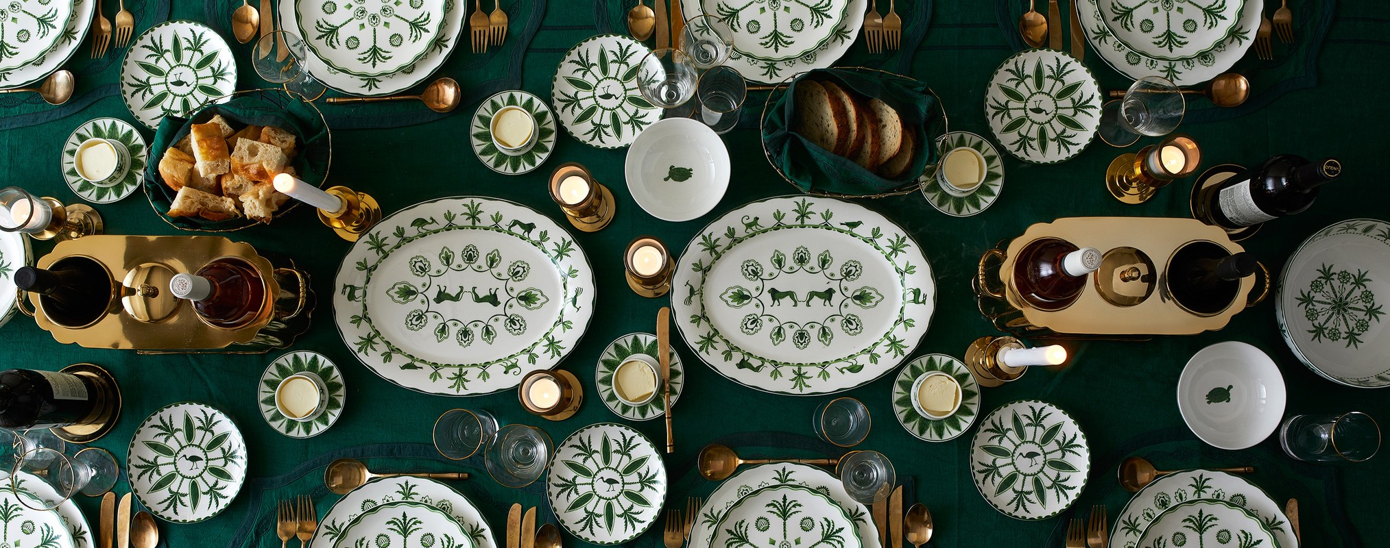Overhead shot of elegantly set dining table, showcasing decorated white china, golden cutlery, candles, and wine, set on a green tablecloth.