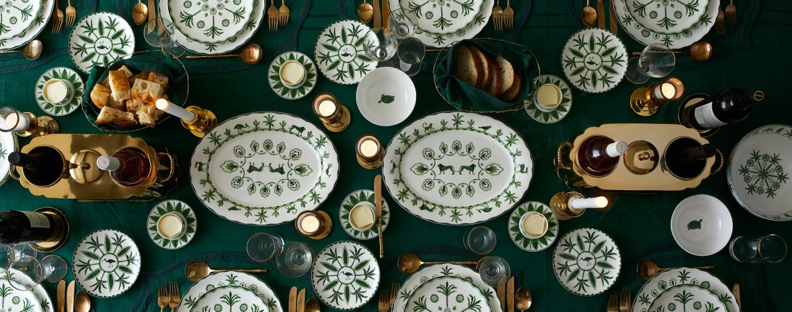 Overhead shot of elegantly set dining table, showcasing decorated white china, golden cutlery, candles, and wine, set on a green tablecloth.