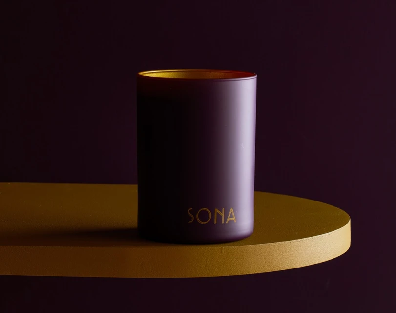 Elegant studio shot of a purple candle with gold accents on a mustard-colored shelf against a rich purple backdrop.
