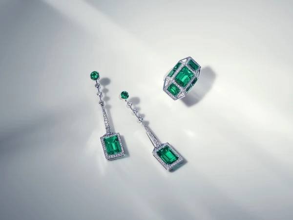 Elegant emerald and diamond jewelry set featuring a ring and drop earrings, arranged on a soft, illuminated white surface. Jewelry photography.