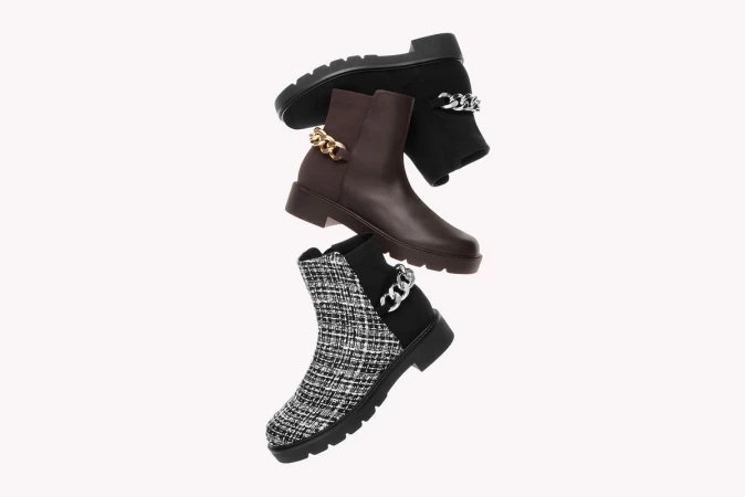Three fashionable ankle boots are stacked against a white backdrop, each with distinctive textures, brown and black tones, and chain embellishments.