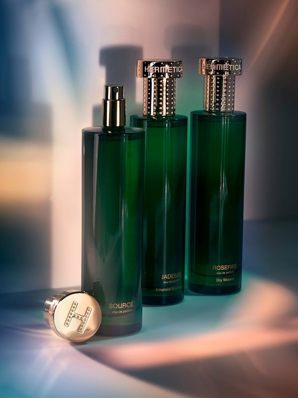 Three green Hermetica perfume bottles stand on a reflective surface with light and shadows. One bottle cap rests in front.