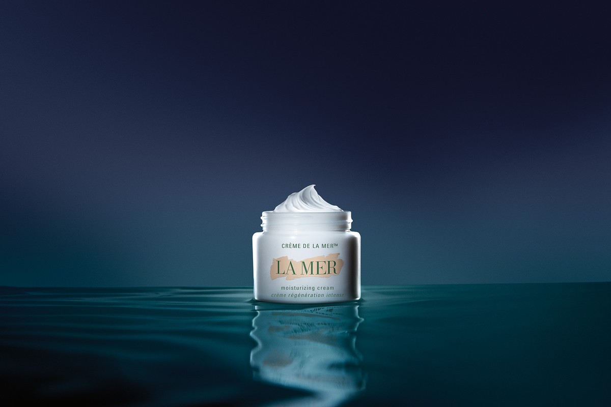 La Mer moisturizing cream jar on a reflective surface with a dark blue background. The focus is on the product, showcasing its texture.