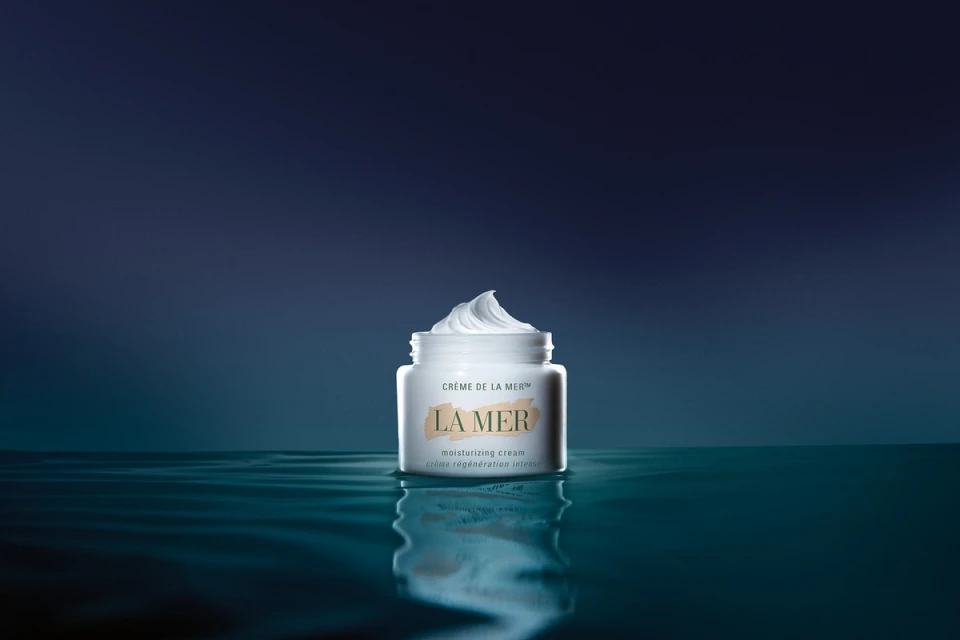 La Mer moisturizing cream jar on a reflective surface with a dark blue background. The focus is on the product, showcasing its texture.