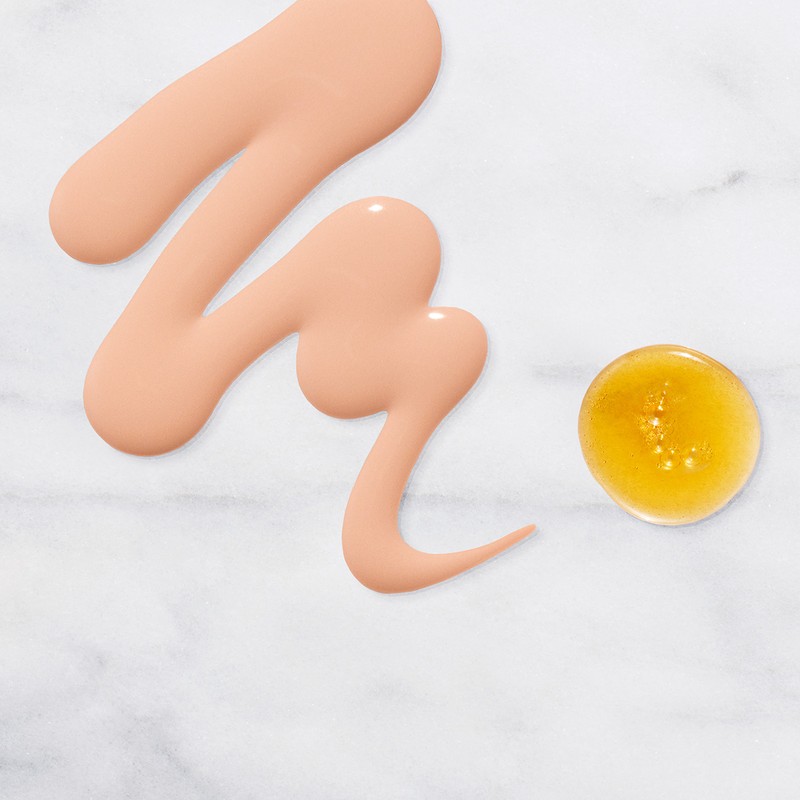 Swatches of skin-toned makeup foundation and yellow serum on a marble surface show product texture and color for beauty applications.